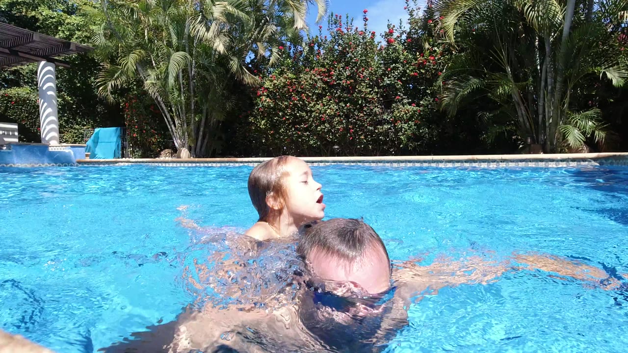 A child surfaces at left while an adult man swims lower in the frame in the Los Cabos pool, with rippled turquoise water filling most of the view.