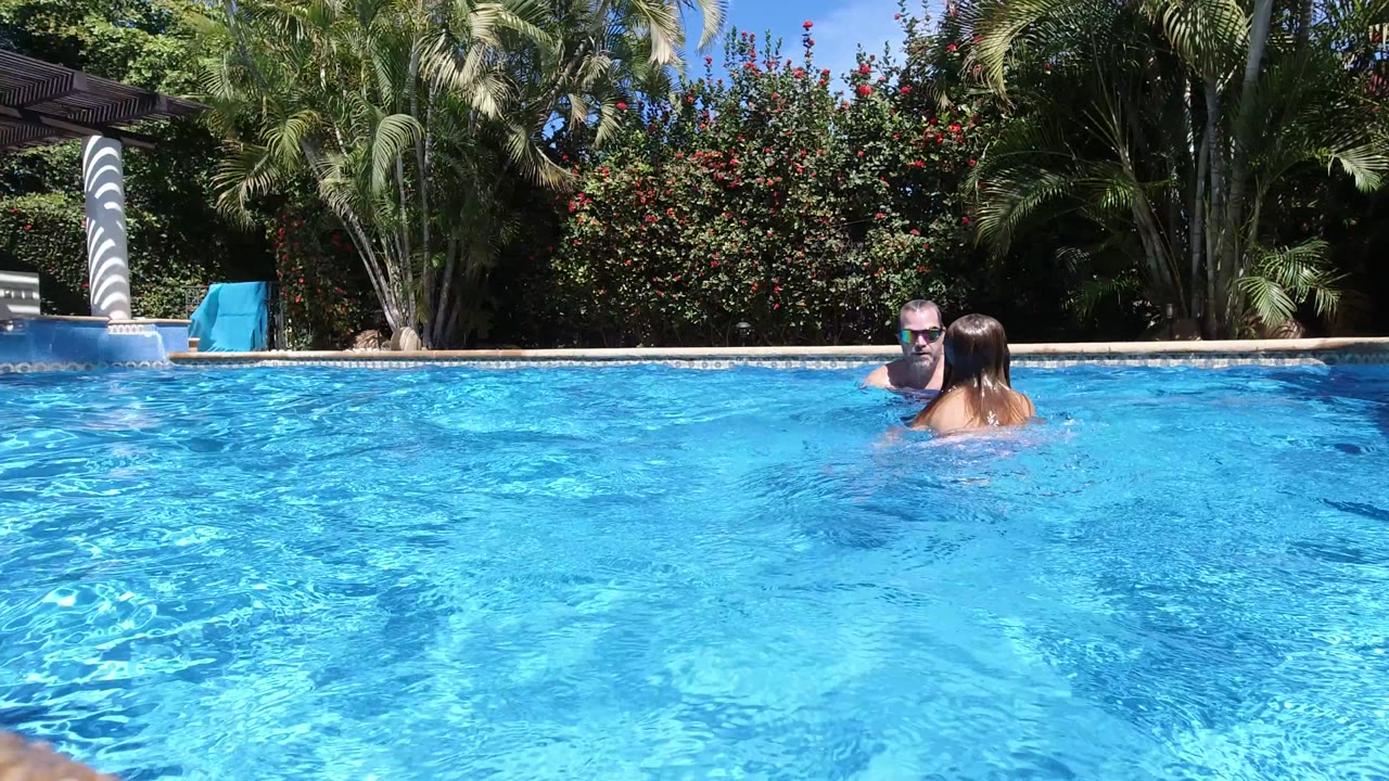 At the start of the Los Cabos clip, a man and young child face each other in a sunlit blue pool with palm trees and flowering shrubs behind them.