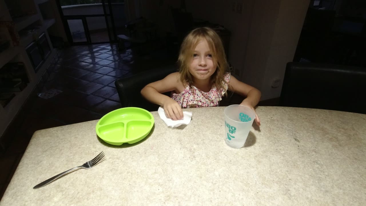 Young blonde girl at a dining table with a green plate, cup, fork, and napkin.