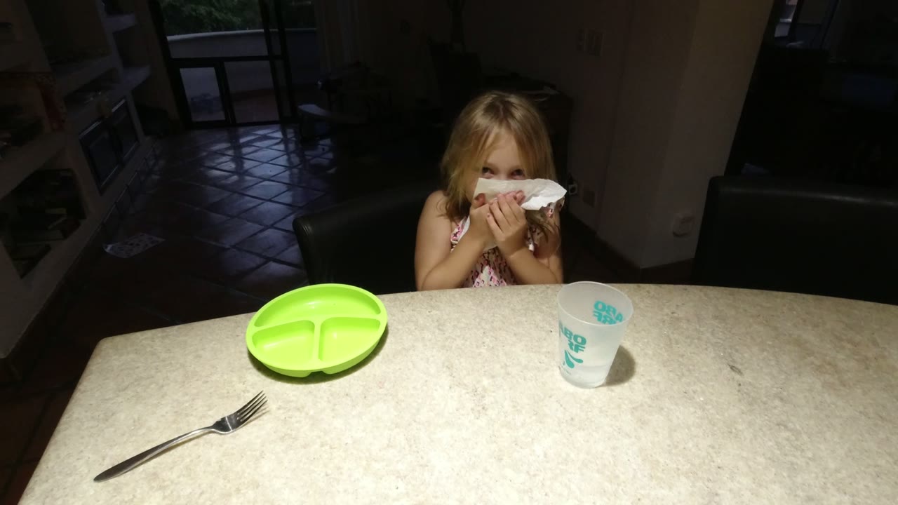 The girl raises a white napkin toward her face at the dining table in Los Cabos, Mexico, while the green plate and cup remain on the tabletop.