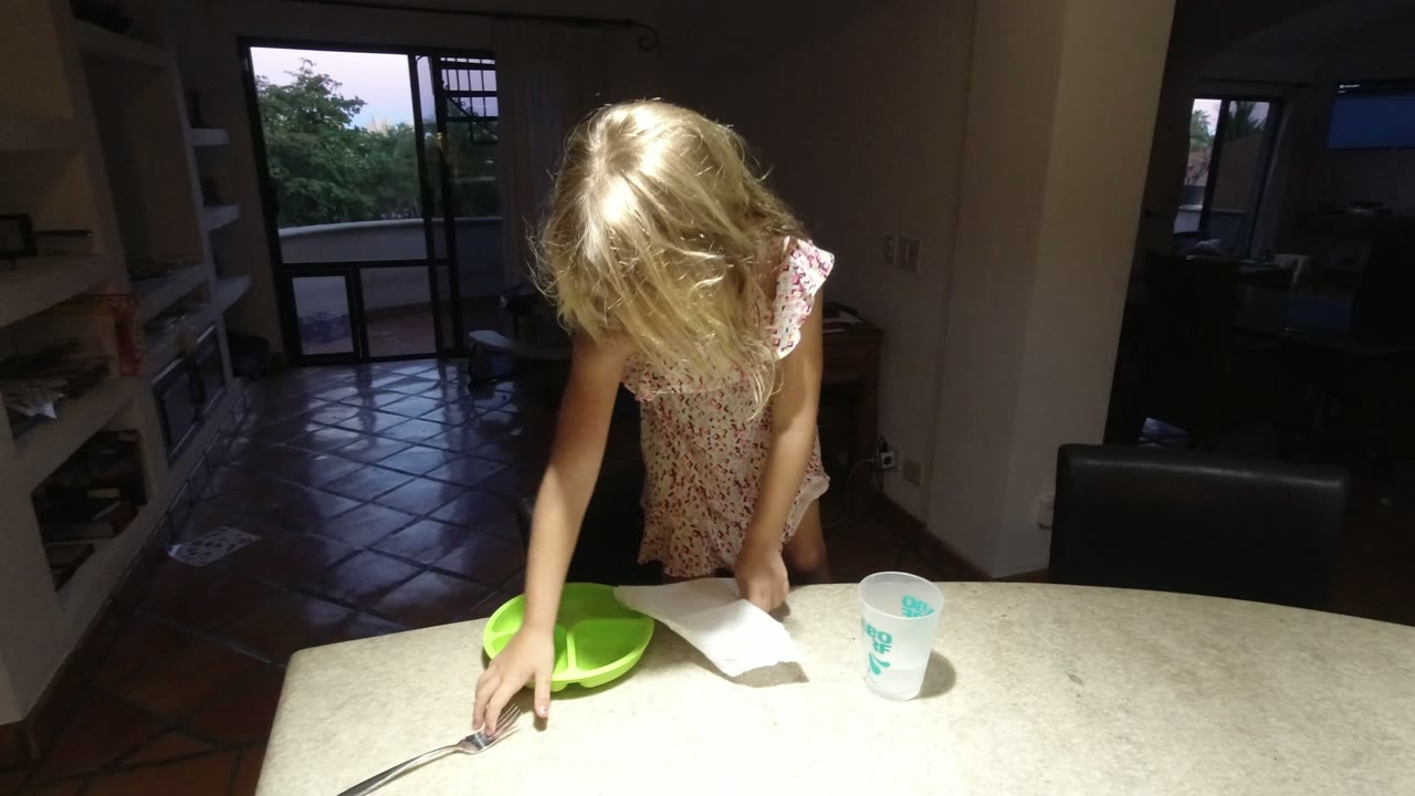 Blonde child at an indoor table with a green plate, fork, napkin, and cup.