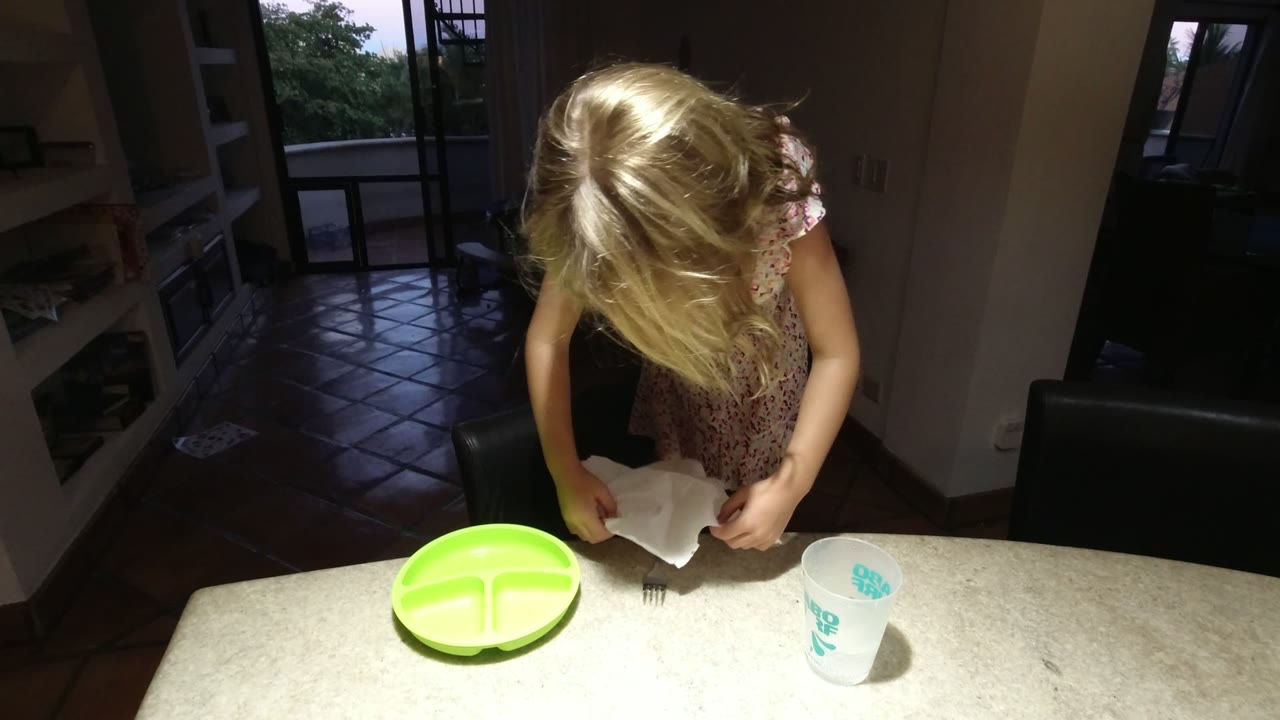 At 0:01 in Los Cabos Mexico, the blonde person bends forward and begins folding the white cloth beside a fork, with the bright green plate centered on the counter.