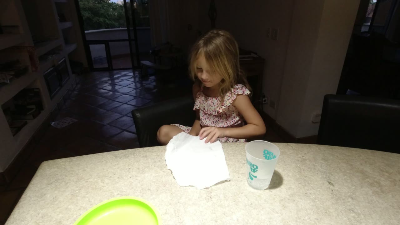 A young girl in Los Cabos sits at a light kitchen table with white paper in front of her, a cup nearby, and a dim open doorway behind her.
