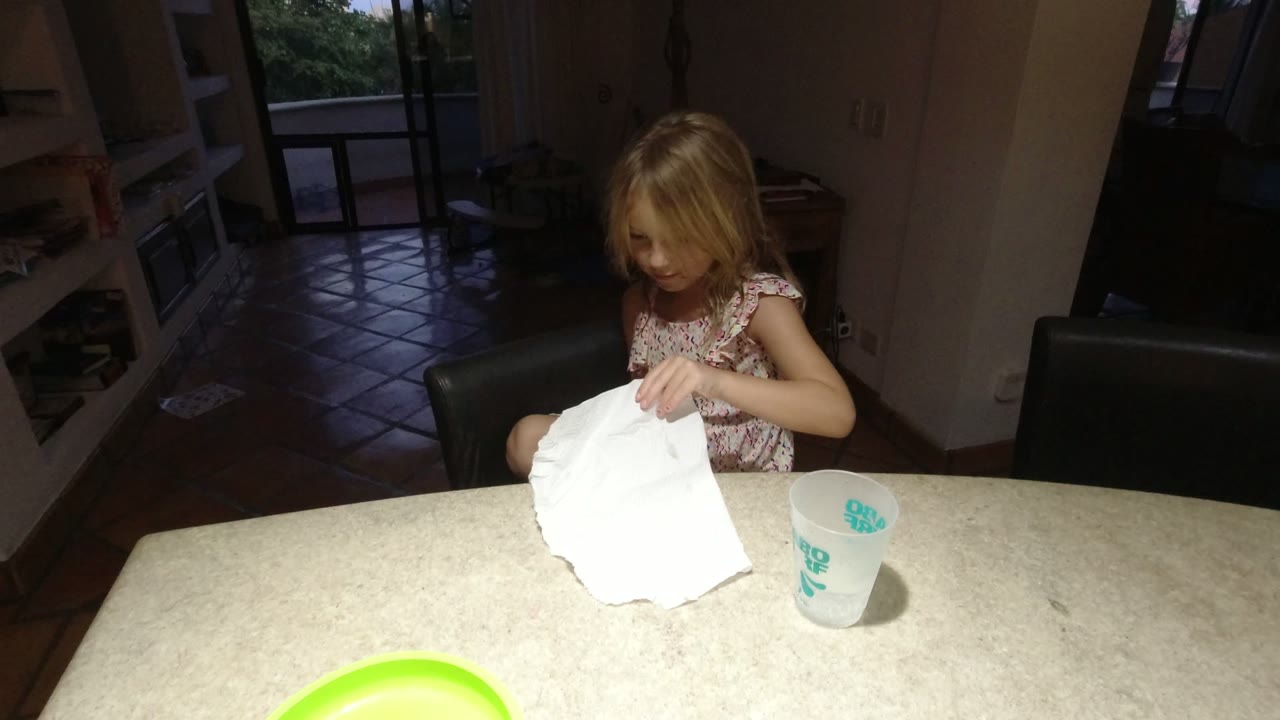 The girl in Los Cabos leans over the table and presses a white paper sheet while a cup sits to her right and the room behind her stays dim.