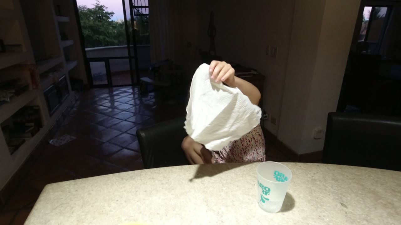 In Los Cabos, Mexico, a young girl sits at a light table with her face mostly covered by a raised white paper towel, with a translucent cup and fork nearby.