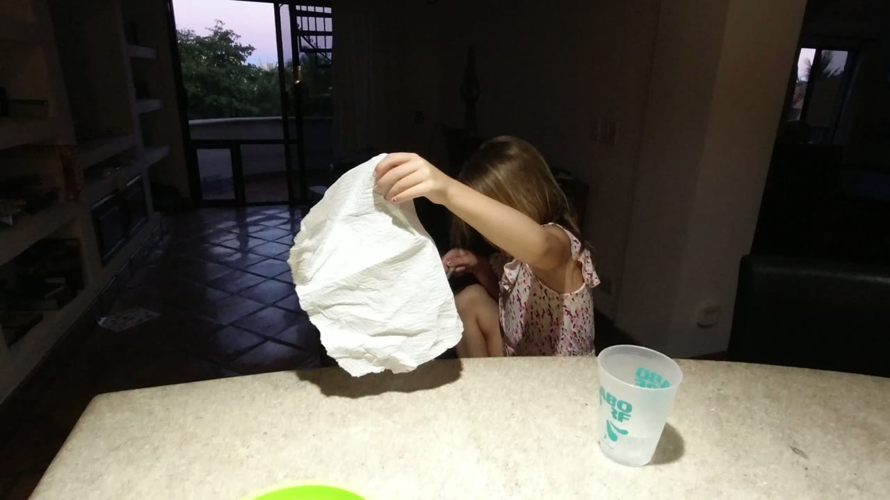 In Los Cabos, Mexico, the young girl holds a crumpled white paper towel high over the tabletop, with one arm and part of her face visible beside a translucent printed cup.