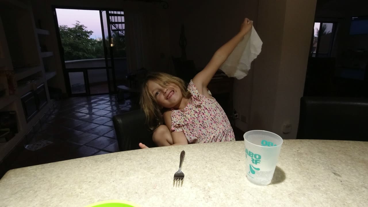 In Los Cabos, Mexico, a young child sits at a light table and raises a white napkin while a fork and plastic cup rest in front of a dim interior.