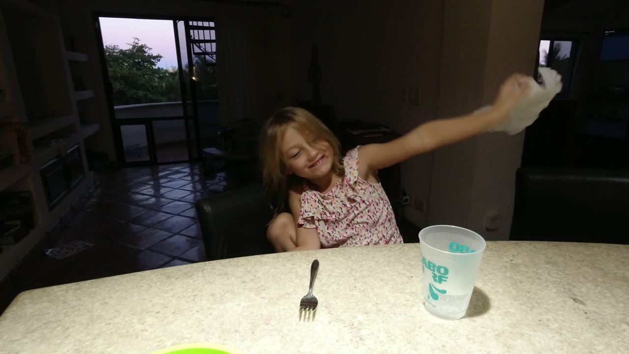 In Los Cabos, Mexico, the child swings the white napkin to one side, with the cup and fork still on the tabletop and the open balcony door visible behind.