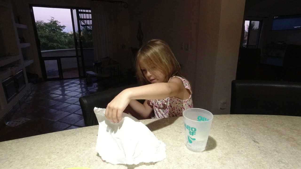 Young girl moves a white tissue on a table beside a translucent cup.
