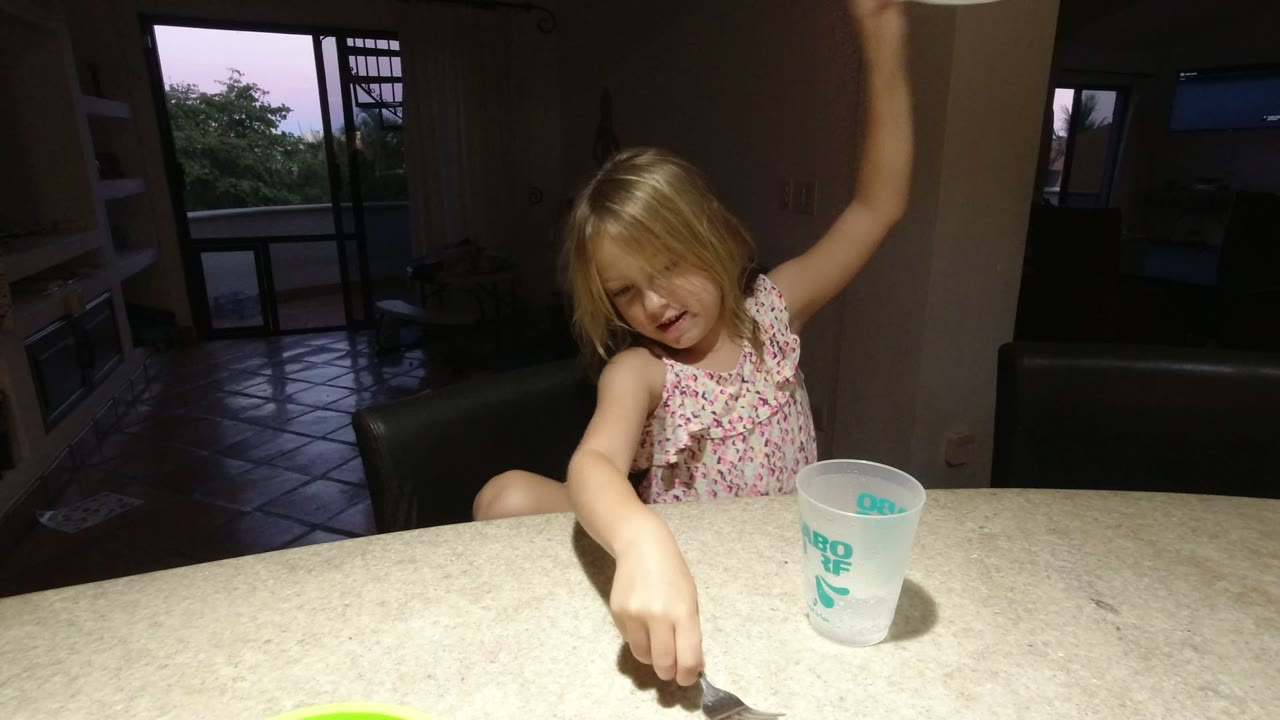 In Los Cabos, Mexico, the child leans over the table with one arm raised and the translucent cup set in front of her, with a dark living area behind.
