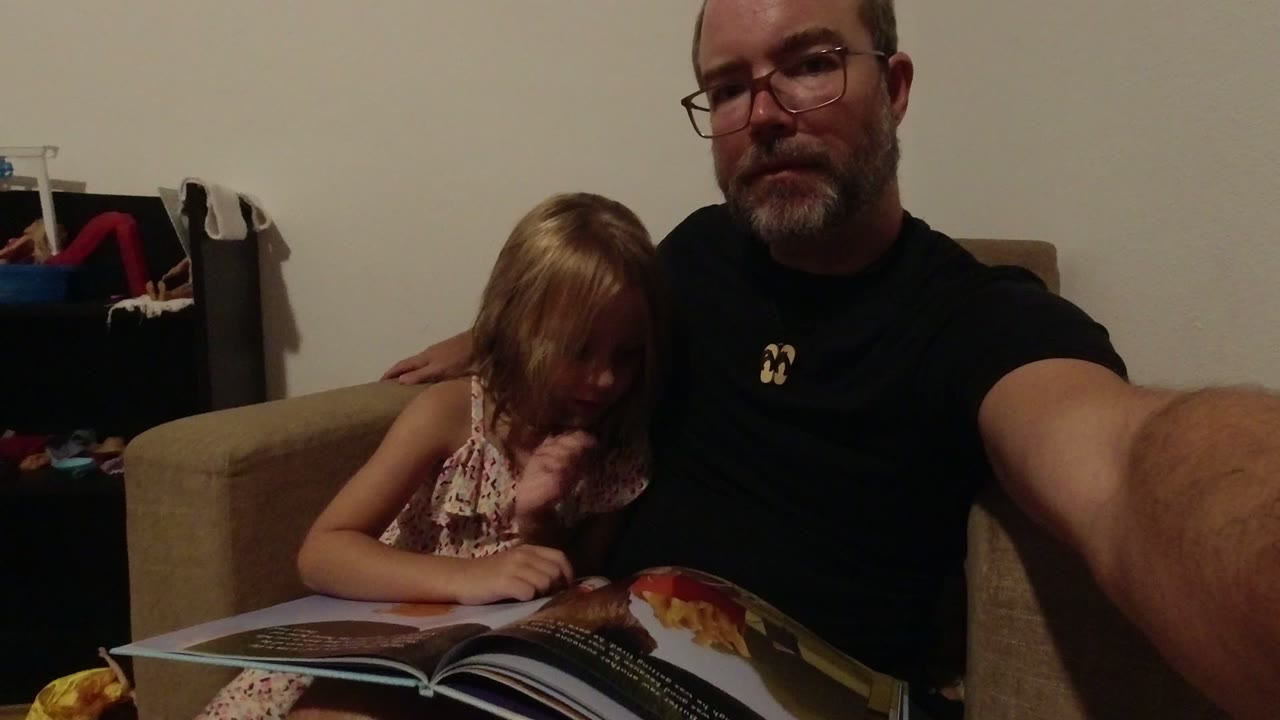 In Los Cabos, Mexico, a father with glasses sits close to a young daughter on a couch as she looks down at an open children's book in warm indoor light.