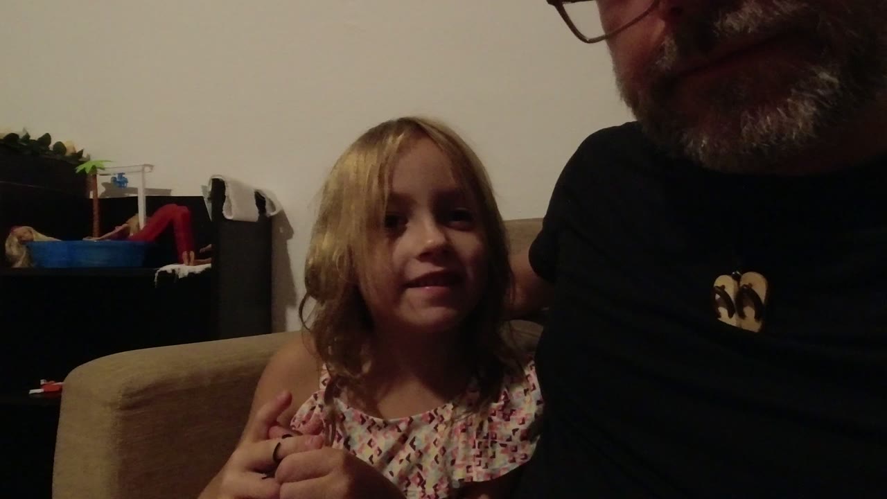 In Los Cabos, Mexico, a young blonde girl sits on a couch beside a tightly cropped adult man in dim indoor light, with shelves and toys behind them.