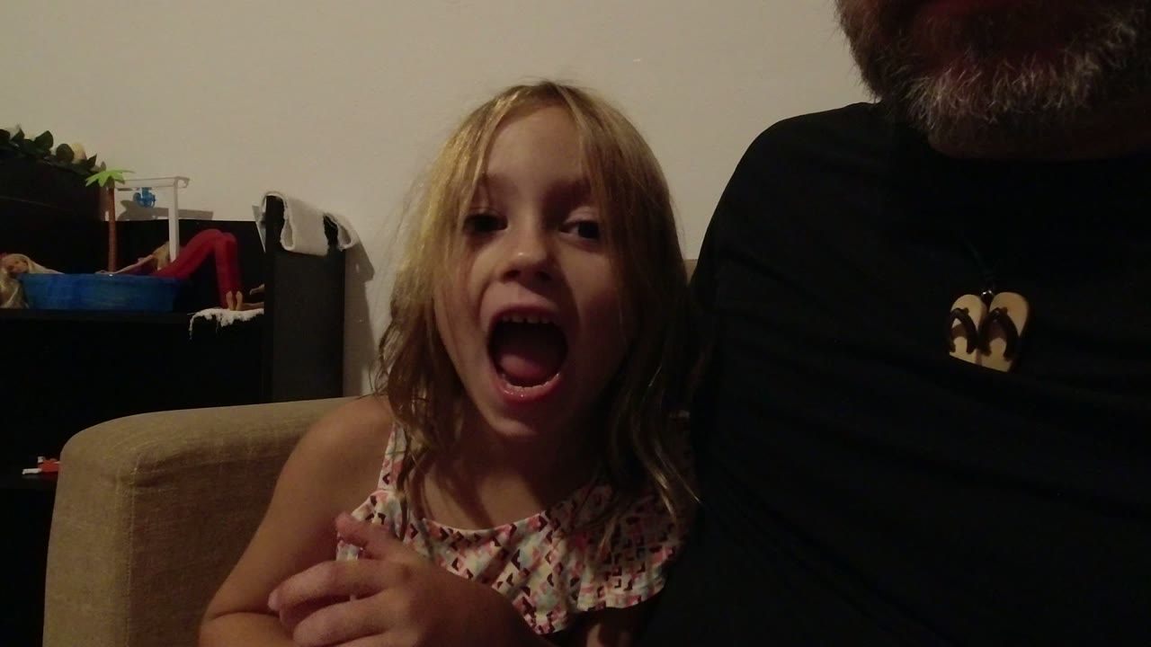 At 0:01, the girl opens her mouth as if speaking or reacting, with the adult's lower face and dark shirt still cropped at the right of the Los Cabos interior frame.