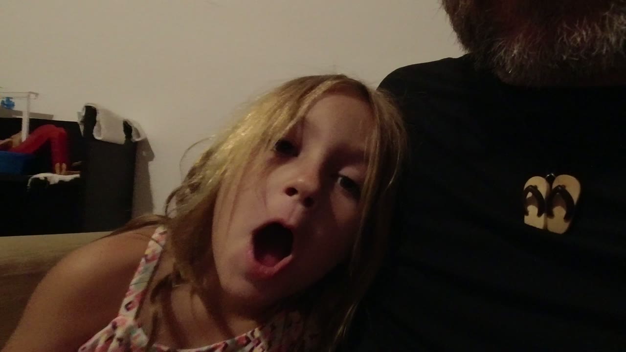 Blonde girl opens her mouth toward the camera beside a cropped adult in a black shirt.