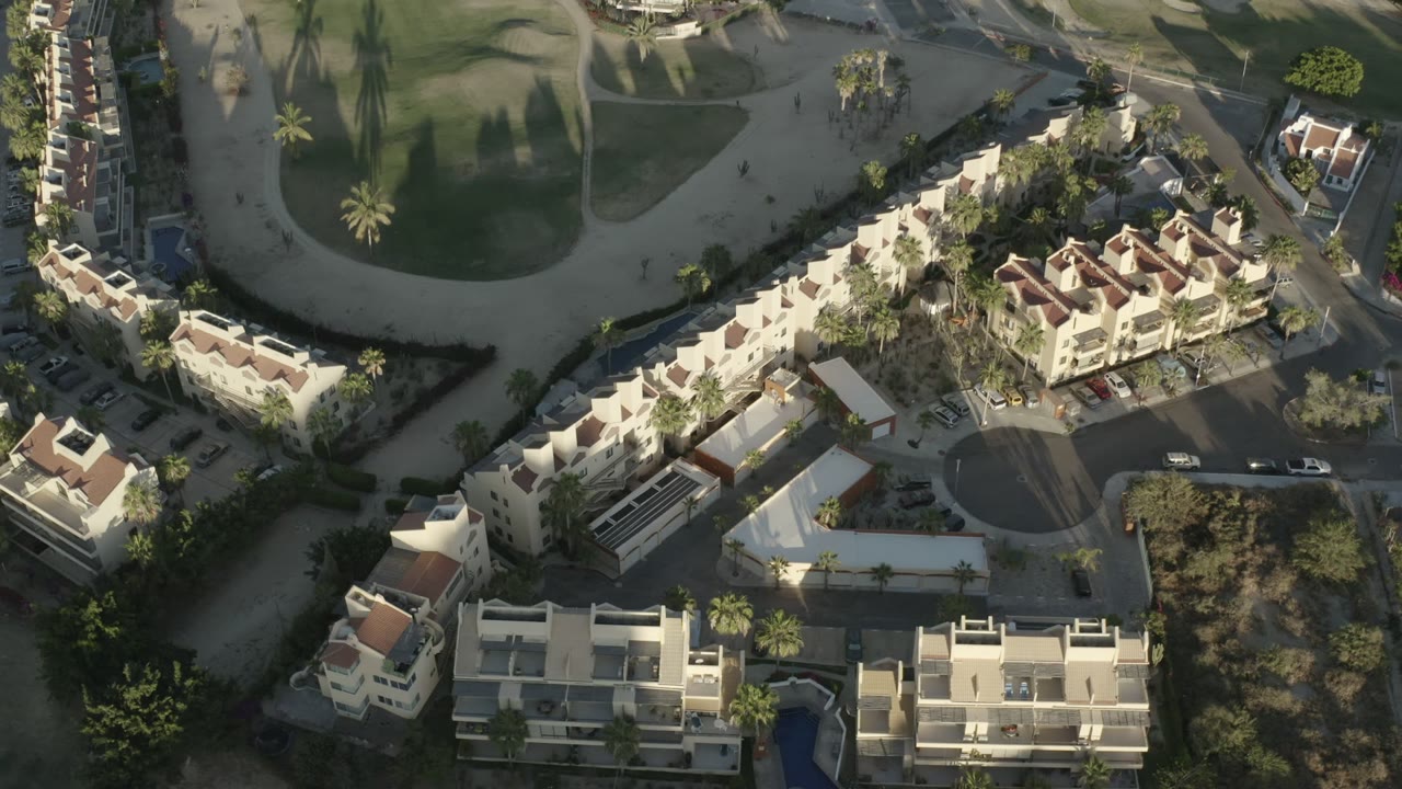 Los Cabos overhead frame with light-colored residential buildings, a curved street, small cars, palm trees, shaded landscaping, and no visible people.