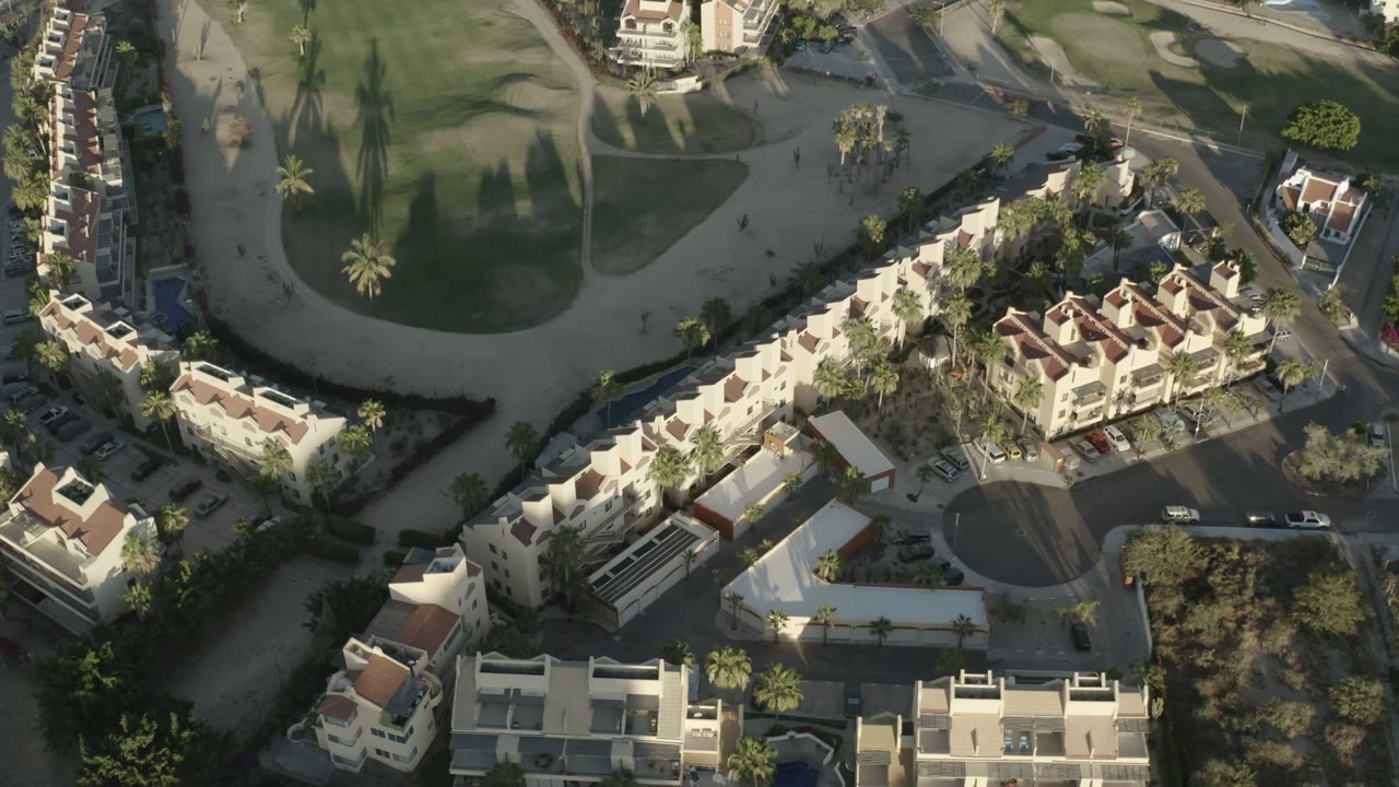 Second frame over Los Cabos condominiums with more of the curved road and parking visible near the buildings, plus golf-course grass and no visible people.