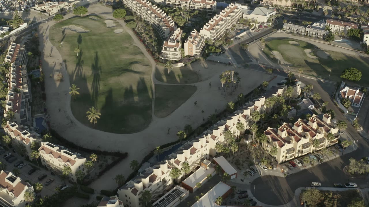 Aerial view in Los Cabos with the golf-course fairway centered, apartment-style homes along both sides, visible roadways, small parked vehicles, and no visible people.