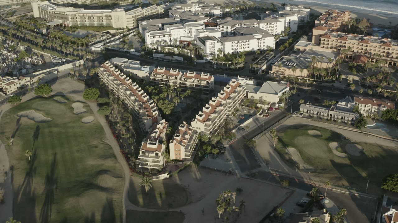 Mid-clip aerial frame in Los Cabos with long shadows across the fairway, clustered palms along roads, residential-style buildings, white structures near the beach, and no visible people.
