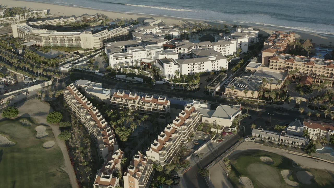 Aerial view over Los Cabos coastal buildings with a golf course in the foreground, a beach and ocean surf beyond, and no visible people.