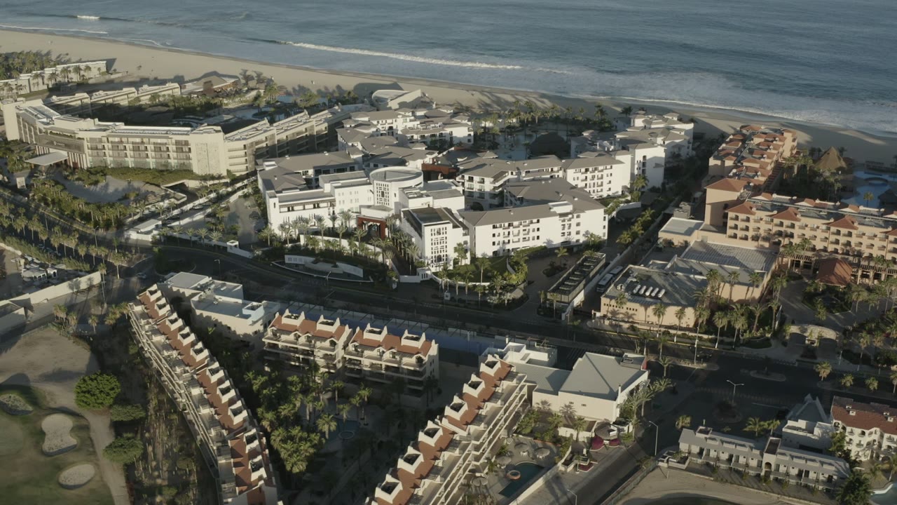 Aerial view in Los Cabos with white resort buildings, rows of terracotta-roofed residences, palm trees, beach sand, ocean water, and no visible people.