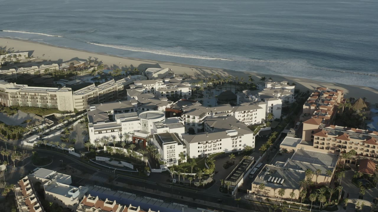 Second Los Cabos aerial frame with the surf line stretching across the top, clustered white buildings in the center, tan-roofed structures to the right, and no visible people.