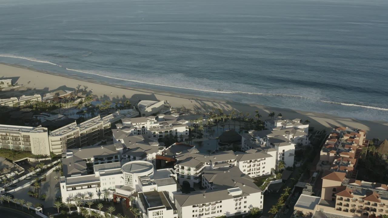 Aerial frame over Los Cabos, Mexico, with blue ocean at the top, a sandy beach below it, white coastal hotel buildings in the foreground, and no visible people.