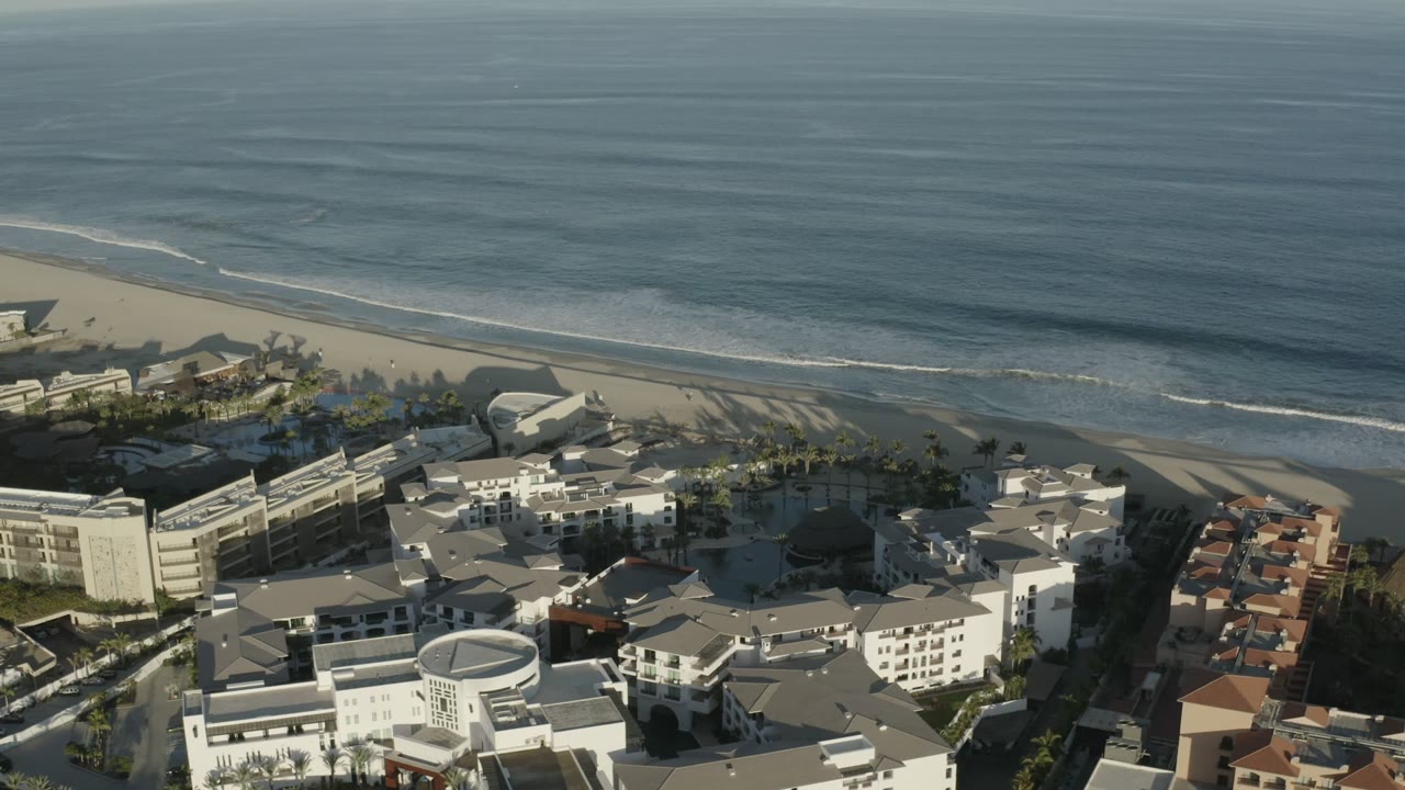 Aerial view in Los Cabos, Mexico, showing gentle surf lines, a wide strip of sand, pools among palm trees, white resort-style buildings, and no visible people.