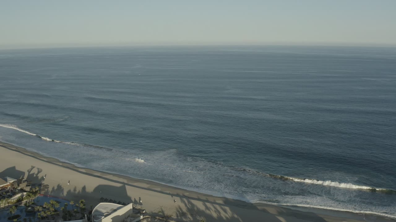Los Cabos aerial coastline view with the surf band slightly farther along the beach, broad blue water in the background, beach in the lower frame, and no visible people.