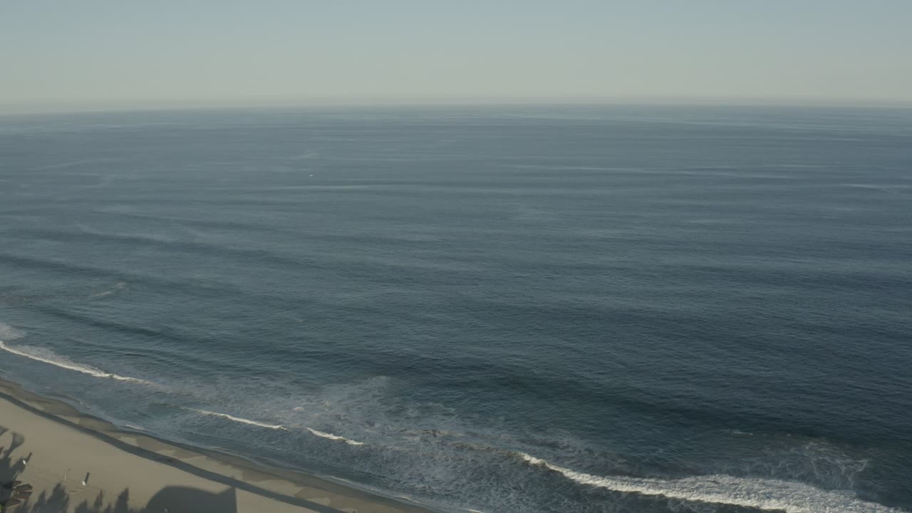 Elevated Los Cabos shoreline view with dark blue water across the frame, a thin white wave line near shore, sandy beach at the bottom, and no visible people.