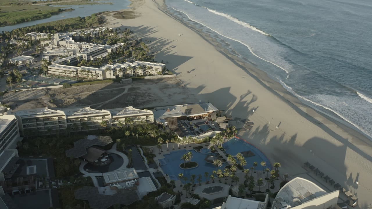High aerial view of surf, sandy beach, pools, palms, and beachfront buildings.
