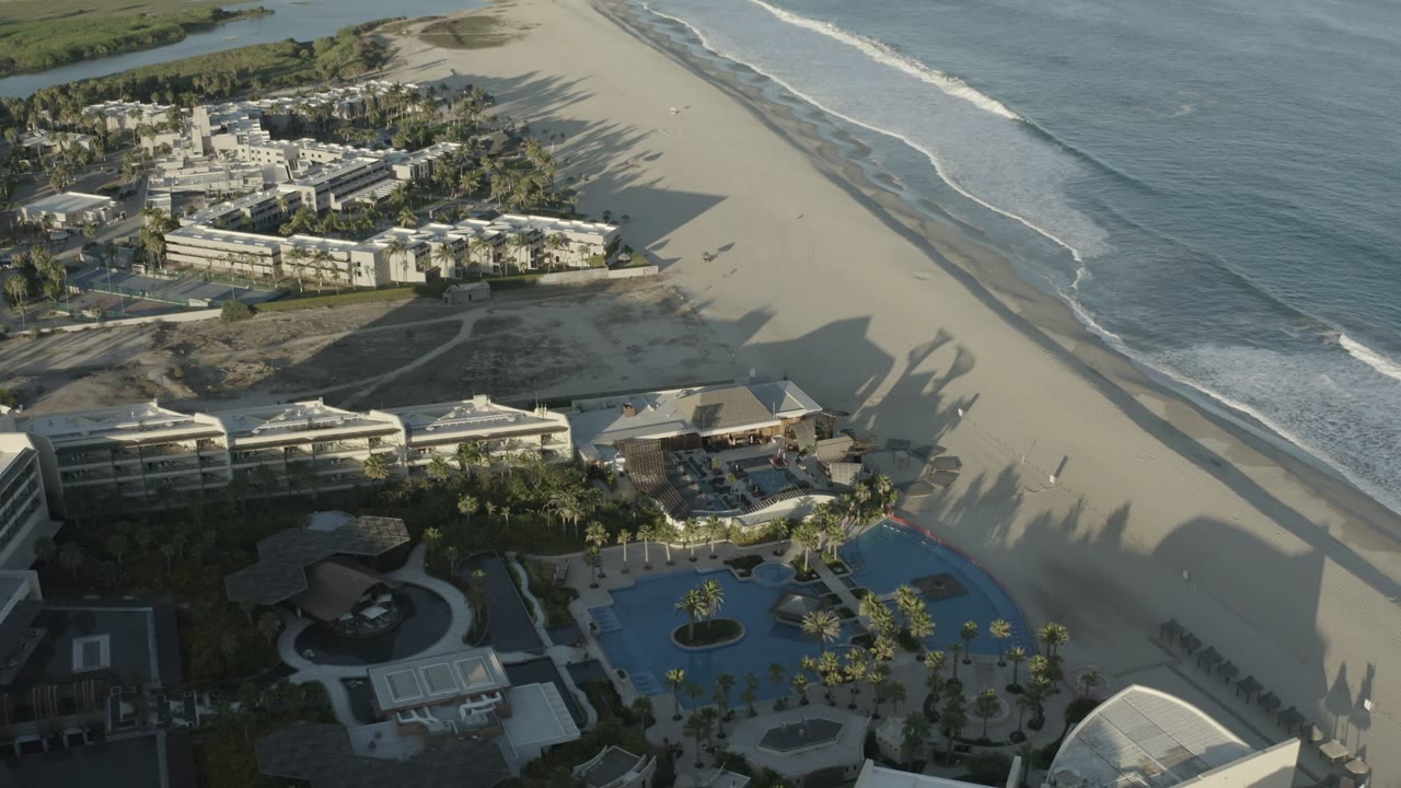 Aerial frame over Los Cabos, Mexico, showing foam lines moving toward the beach, a developed beachfront with pools at the lower edge, and small figures scattered on the sand.