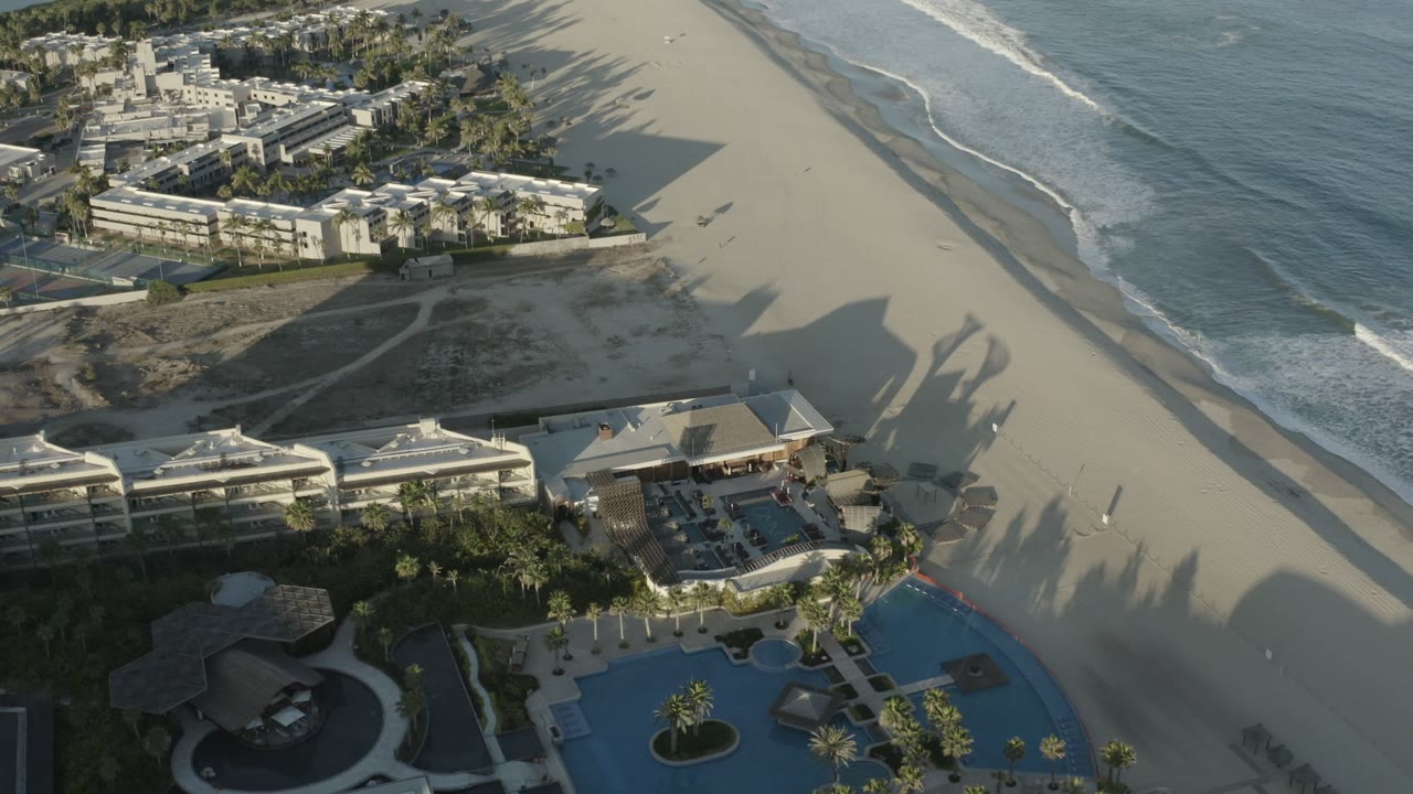 Aerial view of beachfront buildings, pools, sand, surf, and long shadows.