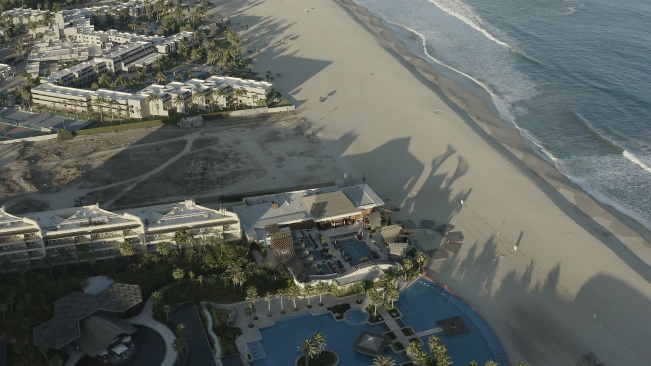 A Los Cabos aerial view shows the ocean line closer to the right edge, beachfront buildings across the lower frame, pool courtyards, long palm shadows, and a few small people on the sand.