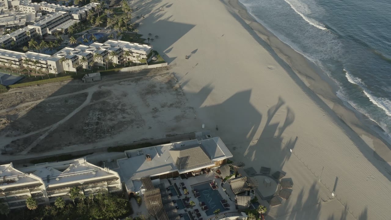 Aerial Los Cabos beach frame with the ocean edge shifted farther right, long building shadows crossing the sand, and tiny beachgoers visible near the open beach.