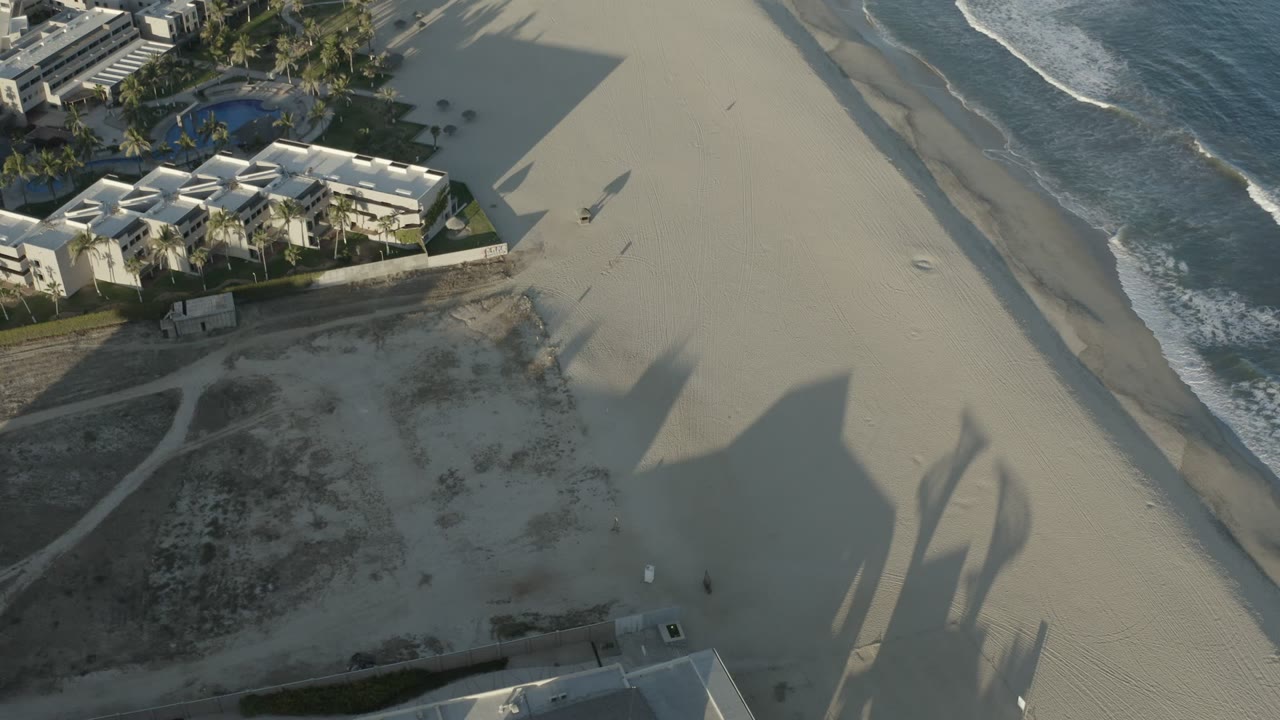 Aerial Los Cabos beach view with the resort buildings shifted toward the upper left, open sand through the middle, breaking waves along the right side, and no visible people.