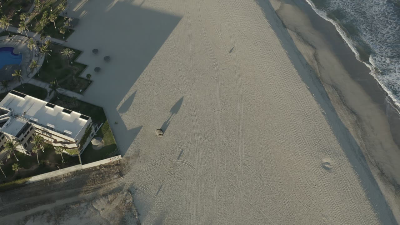 Overhead beach view with sand, surf, palm landscaping, and a white-roofed building.