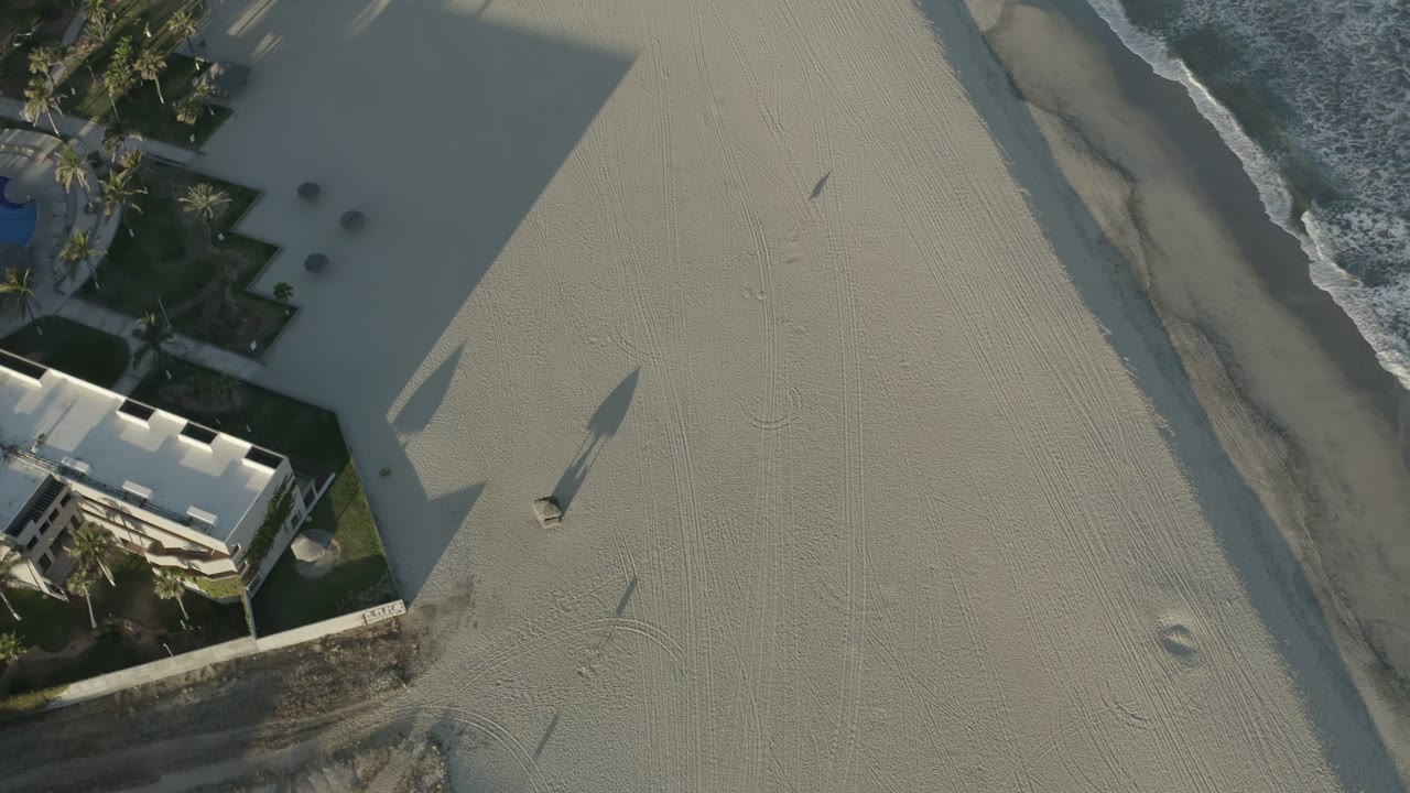 Aerial frame in Los Cabos, Mexico with pale open sand in the center, surf breaking along the edge, palms near a beachfront structure, and no visible people.