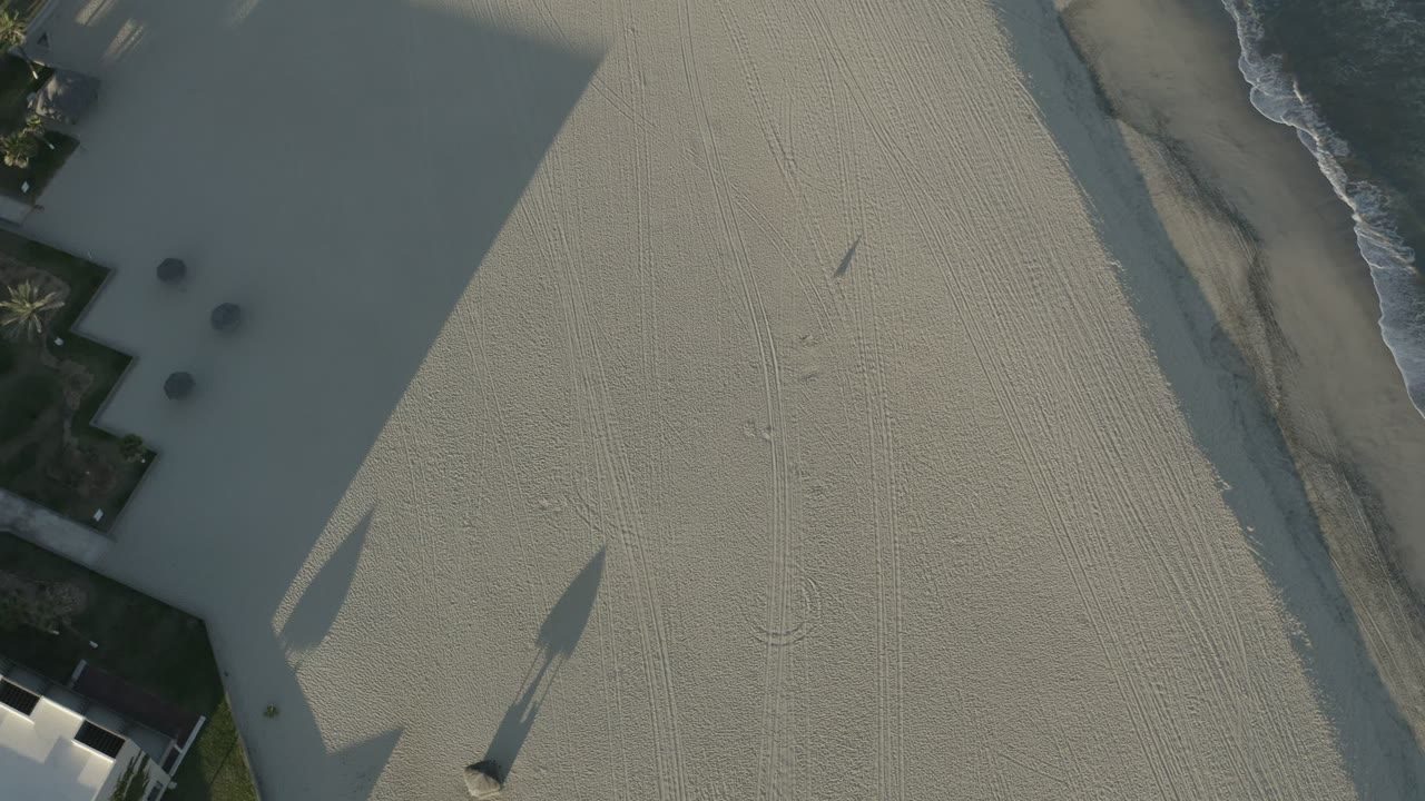 Overhead view of pale ground, dark slab shadows, small round objects, and surf at the edge.