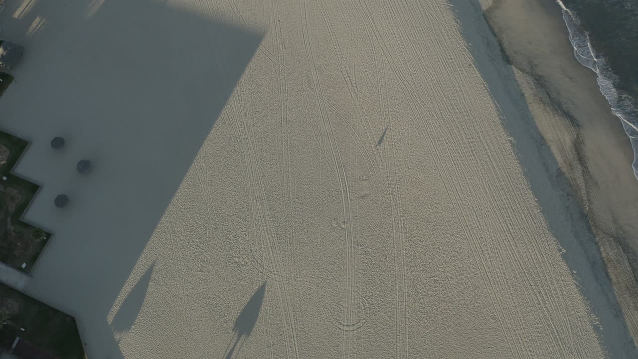 Second overhead frame from Los Cabos, Mexico, with broad dark slab shadows crossing pale textured ground, faint tire or rake marks, surf at the upper edge, and no visible people.