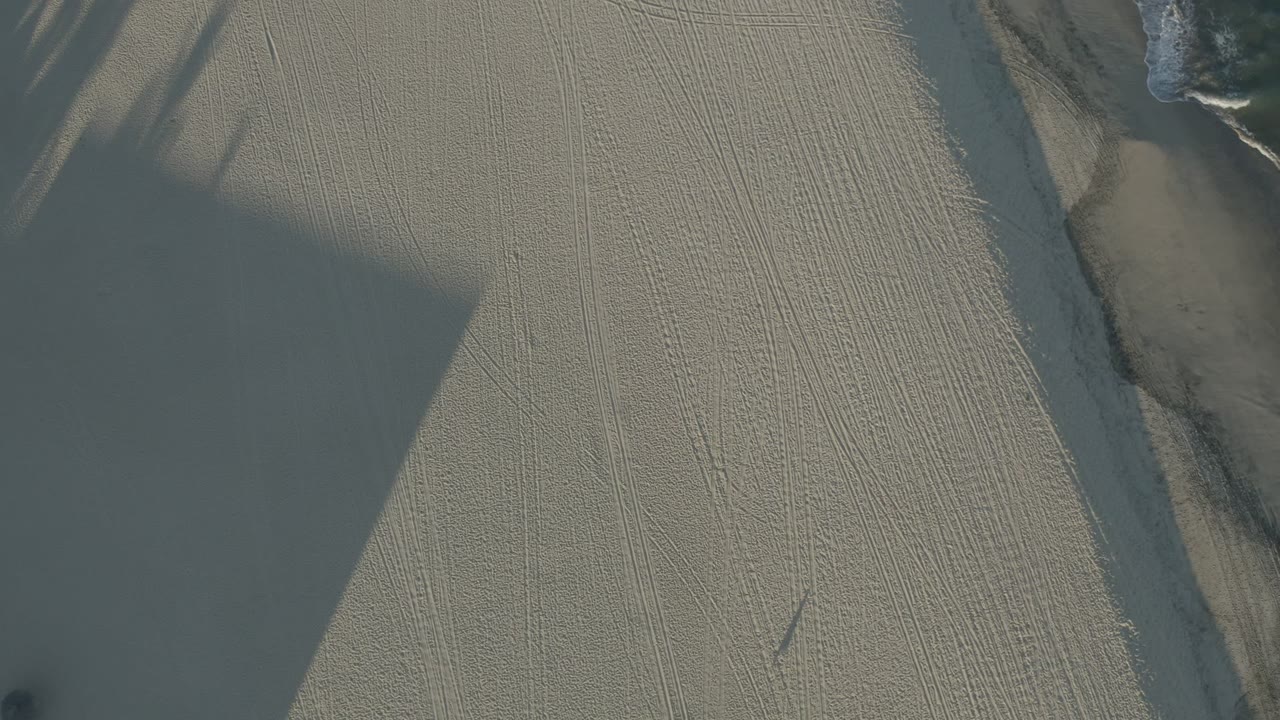Opening overhead frame in Los Cabos, Mexico, showing pale textured sand, angular slab edges, broad shadows, and no visible people.