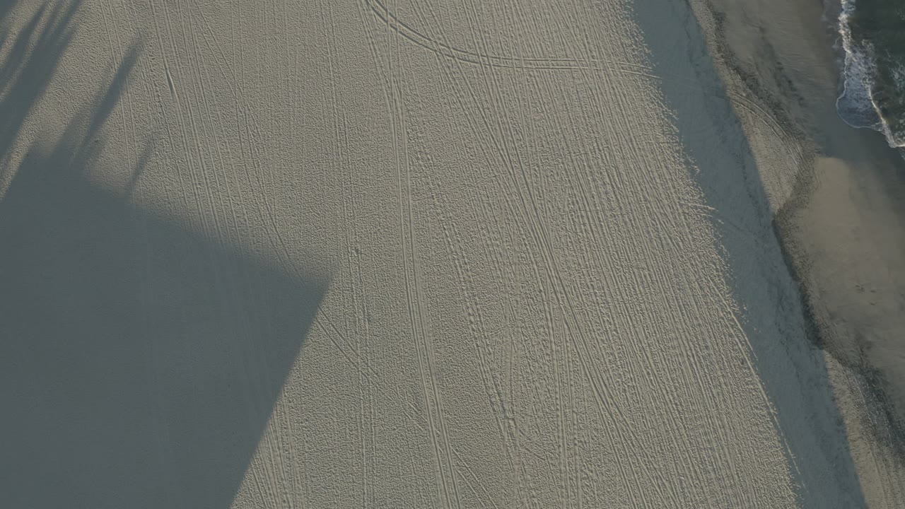 Second overhead frame in Los Cabos, Mexico, with a narrow wet sand channel near the upper area, pale textured ground, a large shadow block, and no visible people.