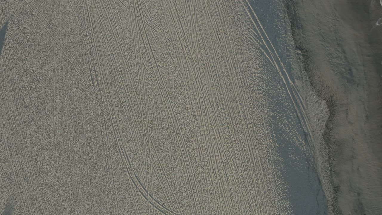 Second Los Cabos overhead frame showing pale sand covered in fine parallel tracks, with a shadowed sand edge crossing the frame and no visible people.