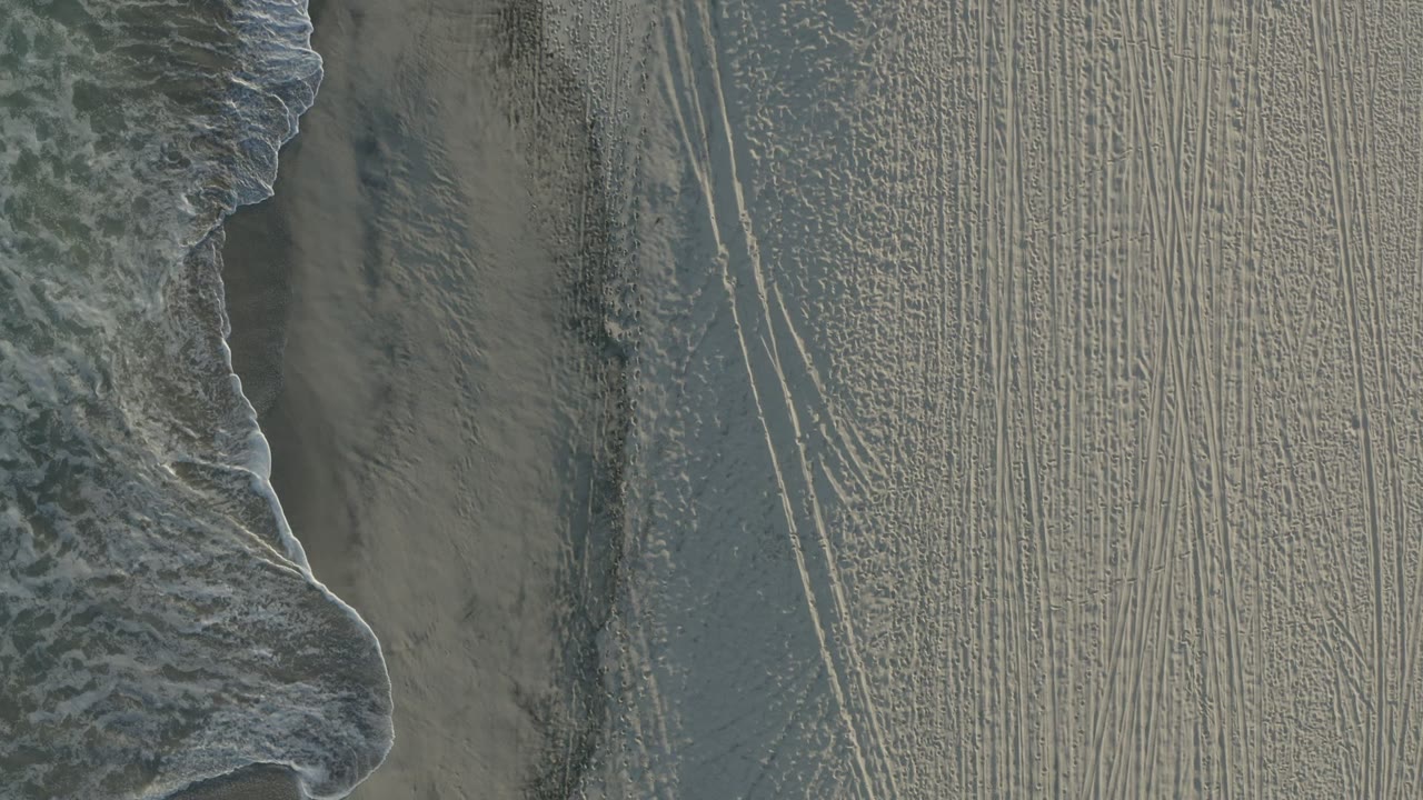 Overhead view of a Los Cabos beach with foamy surf along the left edge, damp sand bands, track marks across the sand, and no visible people.