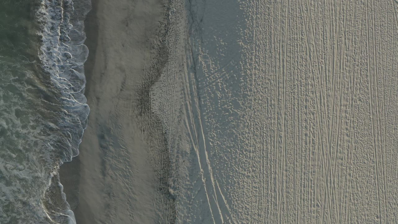 A Los Cabos beach seen from above, with a breaking wave near the center-left, long parallel sand tracks, darker wet sand, and no visible people.