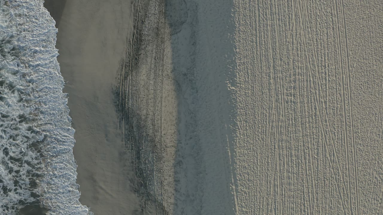 Aerial frame of a thick white foam band beside gray-tan wet sand in Los Cabos, with no visible people.