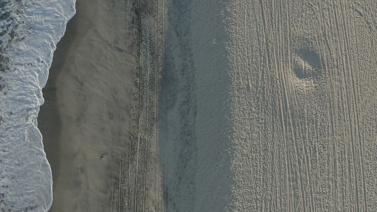 Overhead view of breaking surf and wet sand in Los Cabos, Mexico, with parallel beach tracks and no visible people.