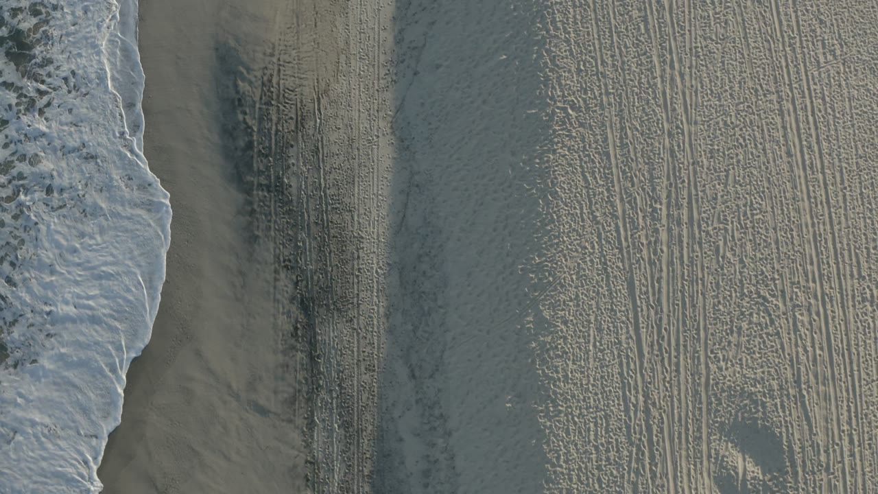 A foamy wave edge runs along textured sand in Los Cabos, Mexico, with a small darker patch in the beach surface and no visible people.
