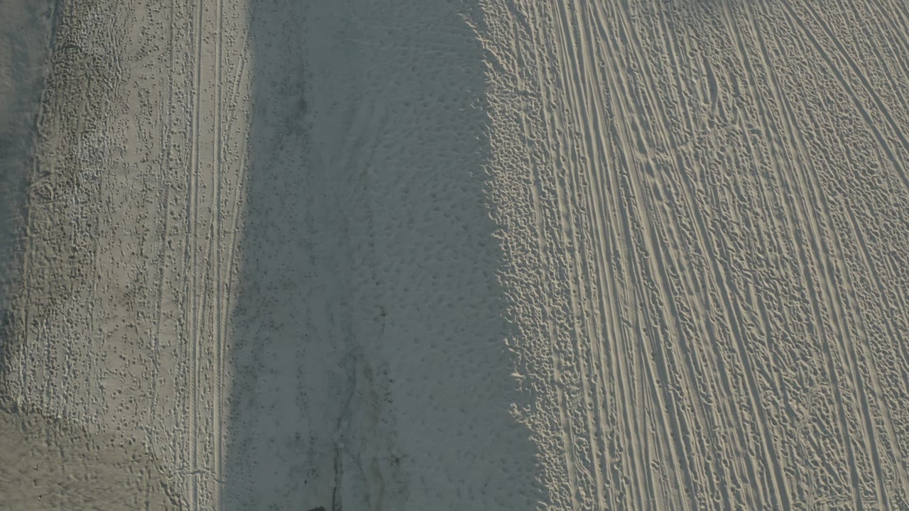 Overhead view of pale sand with parallel tire marks and shadowed ridges.