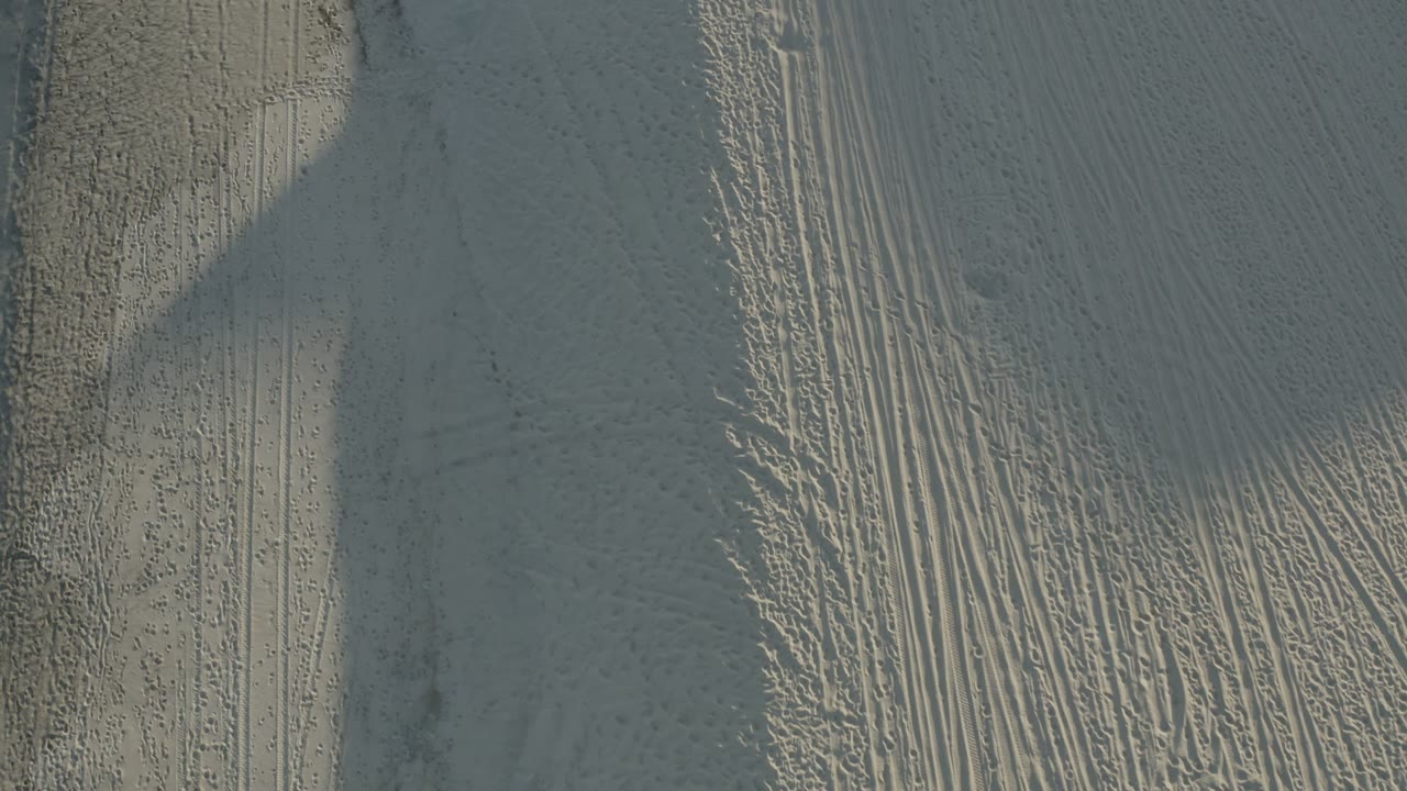 Top-down Los Cabos dune detail with tire tracks running through textured pale sand beside a darker vertical band, with no visible people.