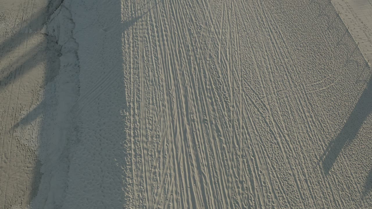 Overhead frame from Los Cabos, Mexico showing pale beach sand, parallel grooming marks, darker shadow bands, and no visible people at the start of the clip.
